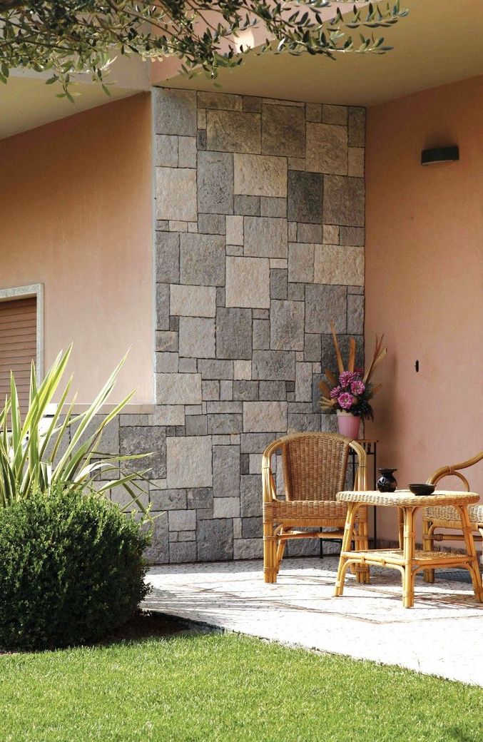 Un patio con un muro con elementi in pietra, due sedie in vimini, un tavolino e un cespuglio verde in una giornata di sole.