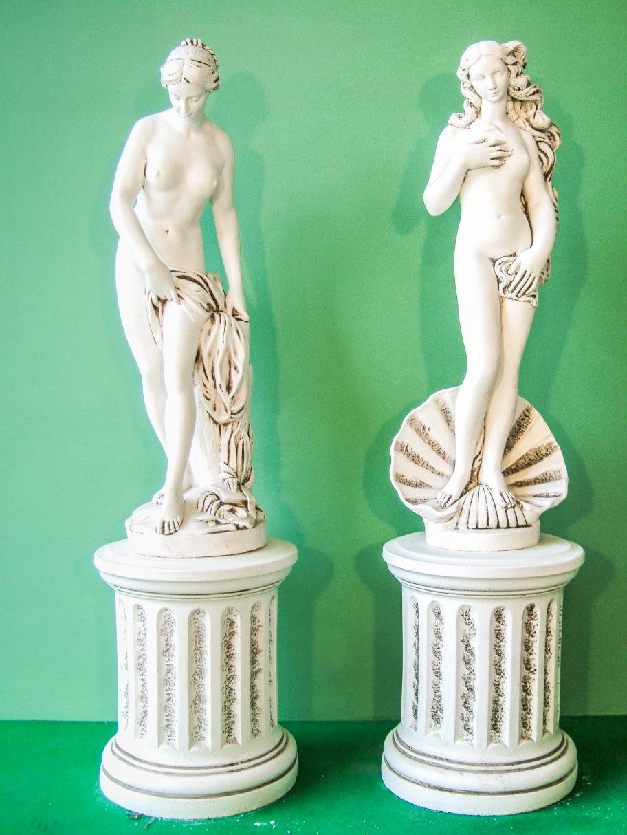 Due statue bianche raffiguranti figure in pose classiche, in piedi su piedistalli scanalati su uno sfondo verde.