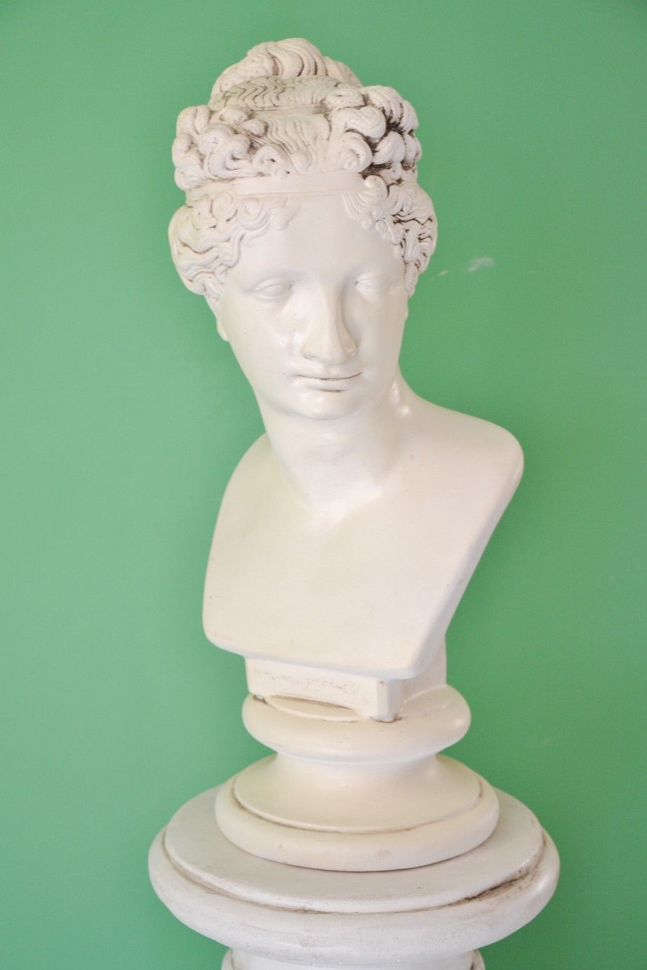 Un busto in pietra bianca raffigurante una figura classica con capelli ricci, su uno sfondo verde uniforme.