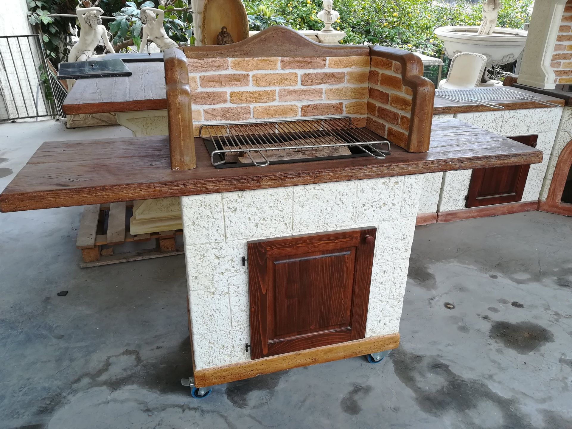 Un barbecue portatile da esterno con retro a mattoni, piano di lavoro in legno e anta scura su base bianca maculata.