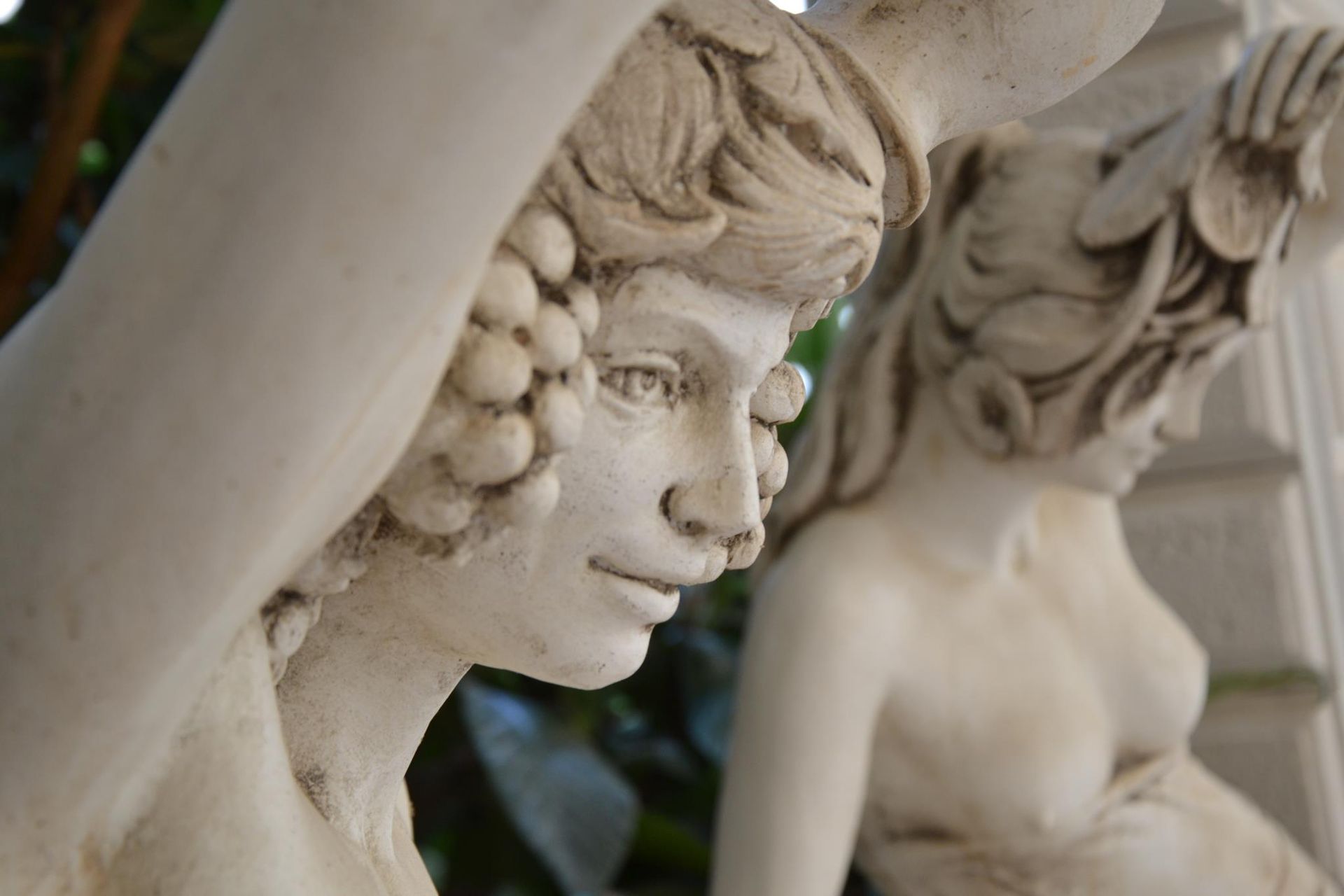 Primo piano di sculture in pietra raffiguranti figure classiche adornate con grappoli d'uva, sullo sfondo di fogliame verde.