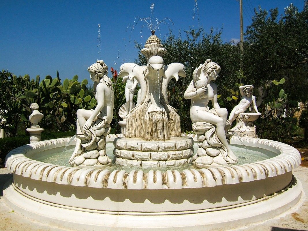 Una fontana in pietra presenta al centro delle sculture raffiguranti dei delfini, circondate da statue sedute.