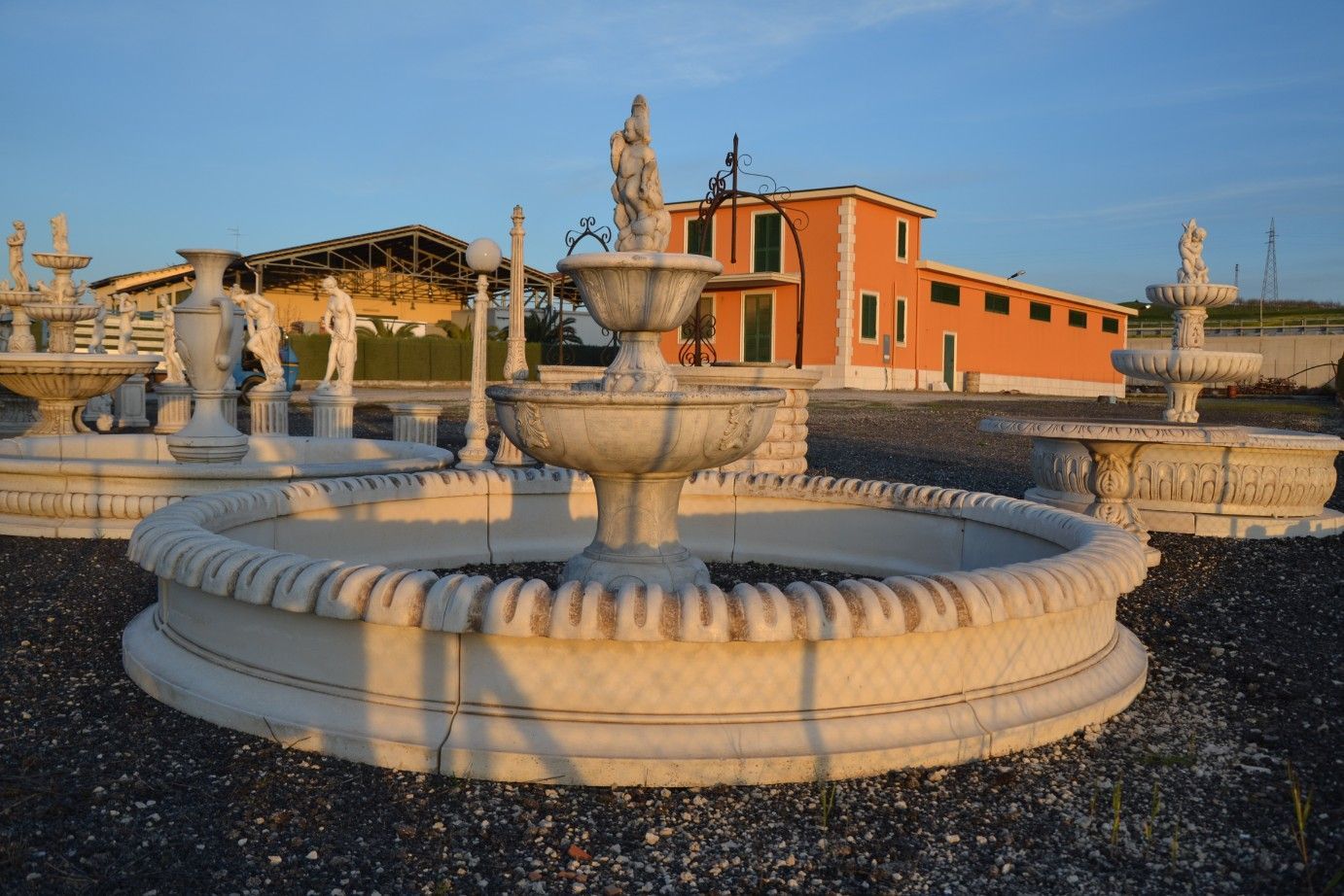 Una fontana di pietra con una statua al centro si trova su un terreno ghiaioso di fronte a un edificio arancione.