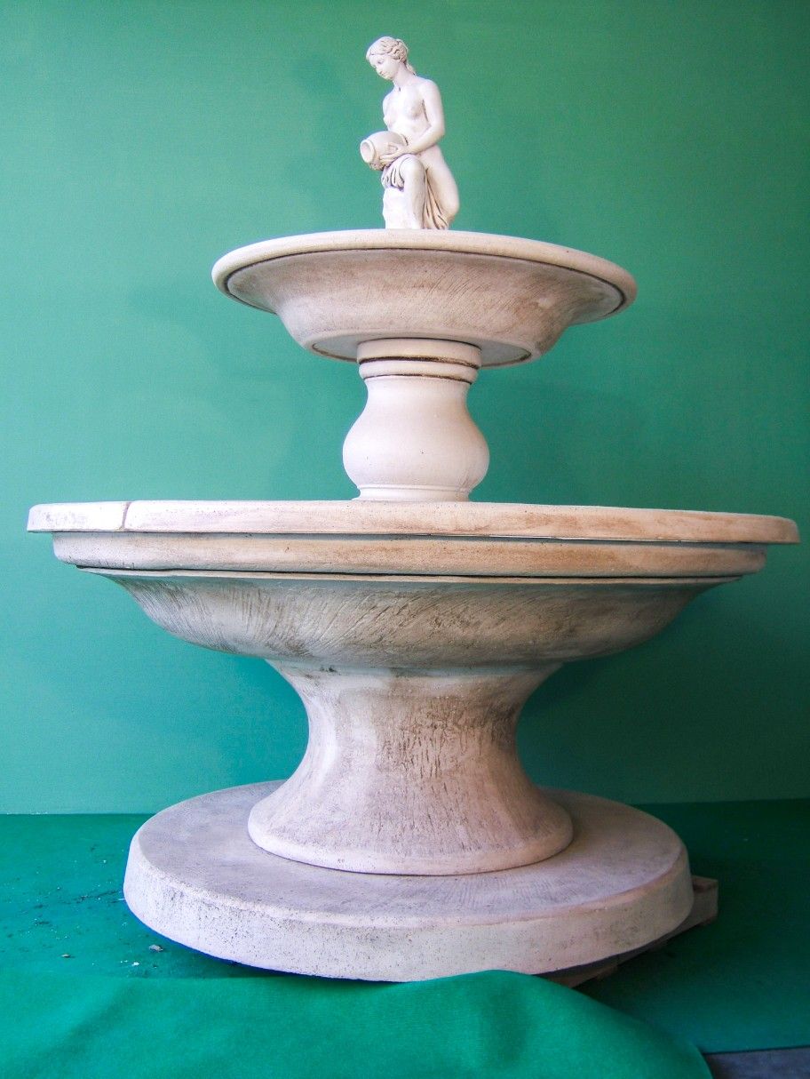 Fontana da giardino in pietra chiara a due livelli, con una piccola statua in cima e posta su uno sfondo verde semplice.