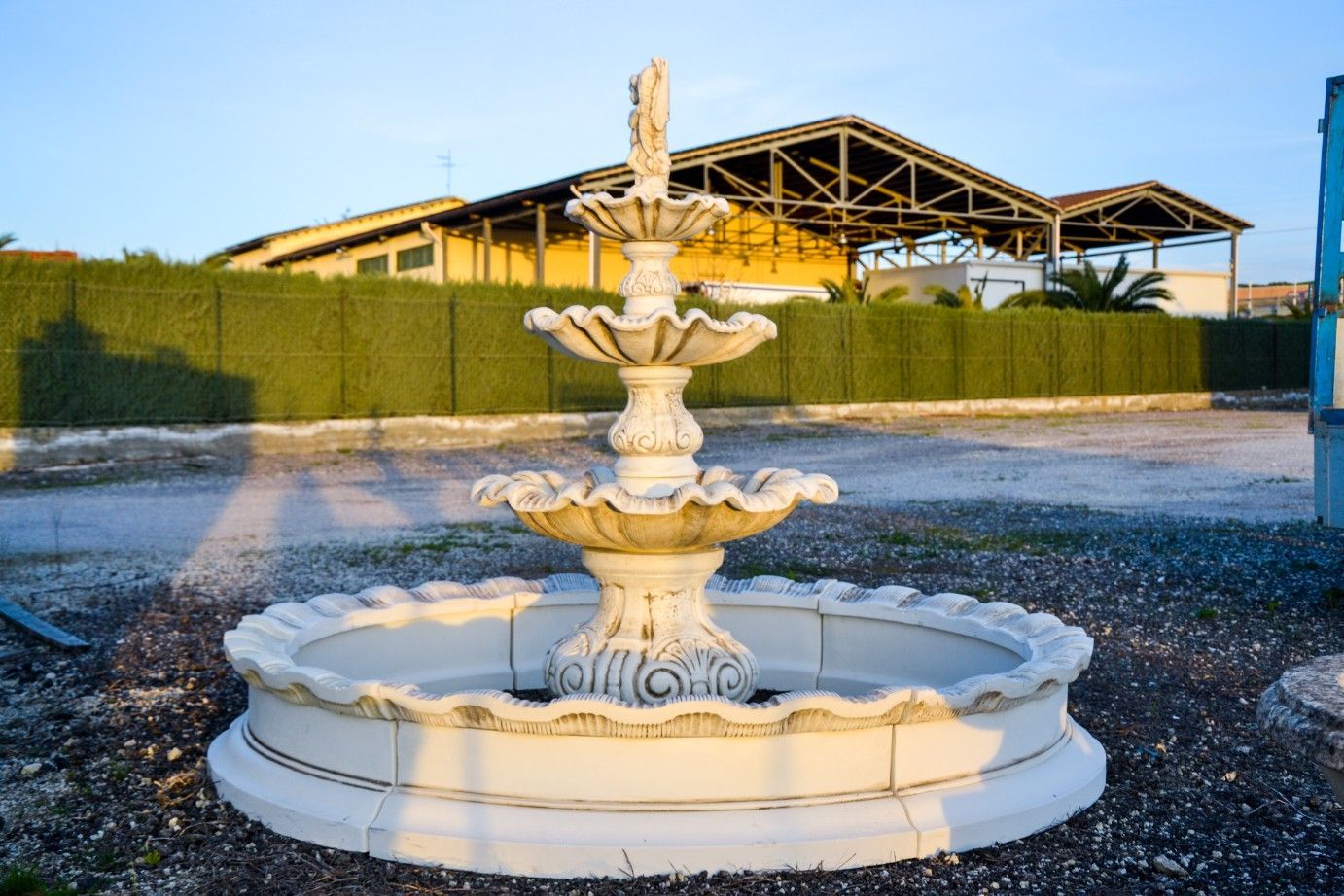 Una fontana in pietra bianca a tre livelli si trova in un terreno ghiaioso di fronte a un'alta siepe verde e a un edificio.
