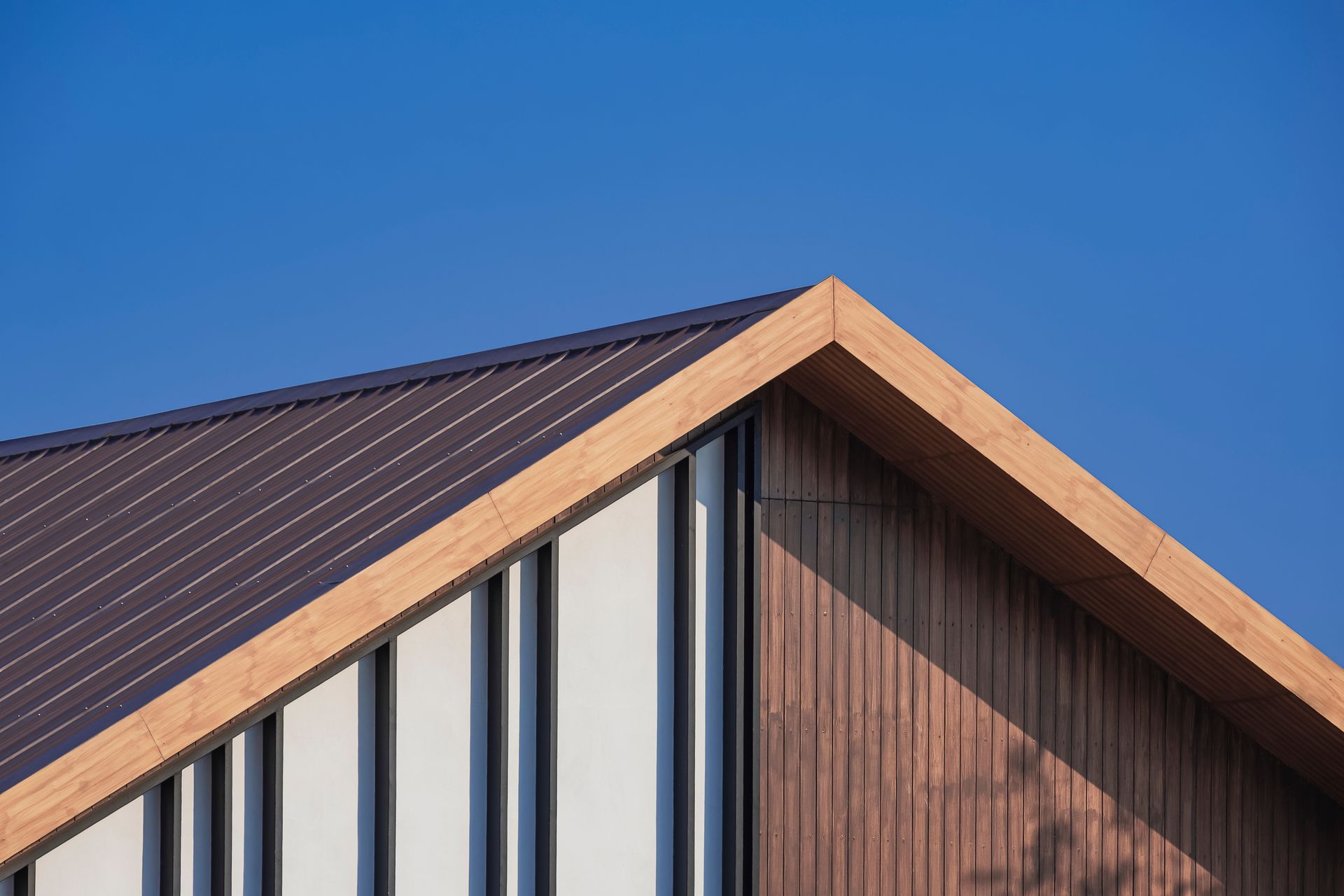 Modern house roof with dark metal panels, wood trim, and vertical white siding against a blue sky