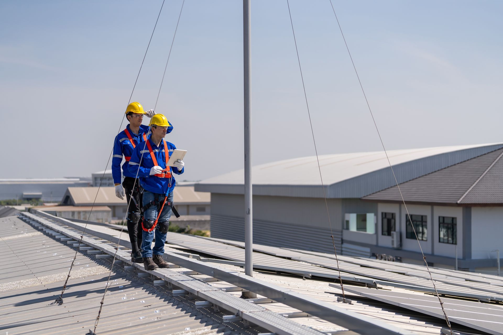Two workers in safety gear inspect rooftop solar panels with harnesses on a commercial building.