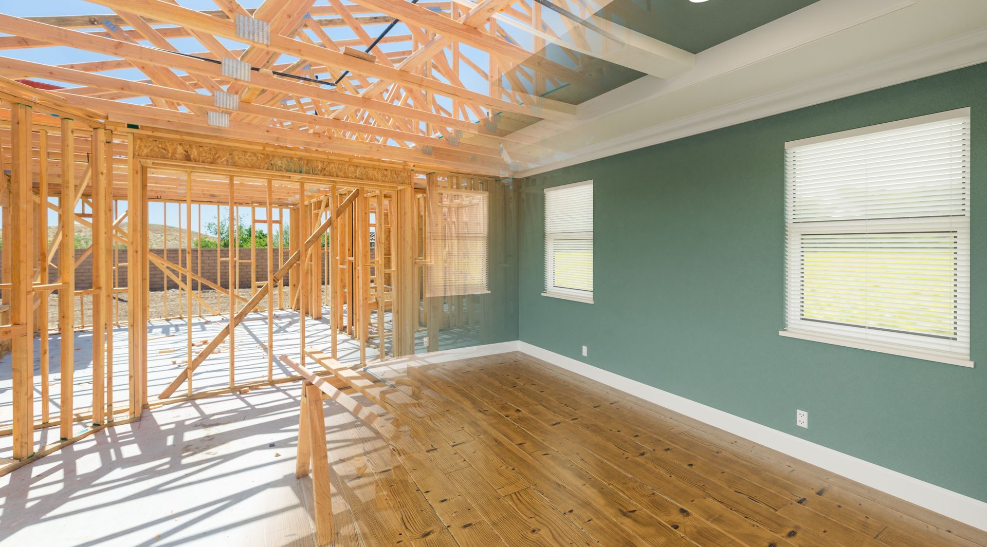 Sunlit room under construction with exposed wood framing, teal walls, and hardwood floor