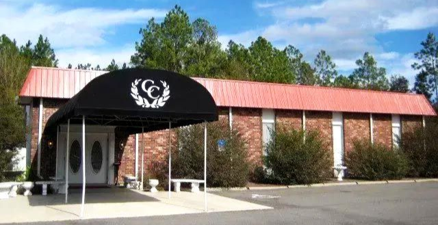 Brick building with a red roof and black awning at the entrance, surrounded by trees under a blue sky