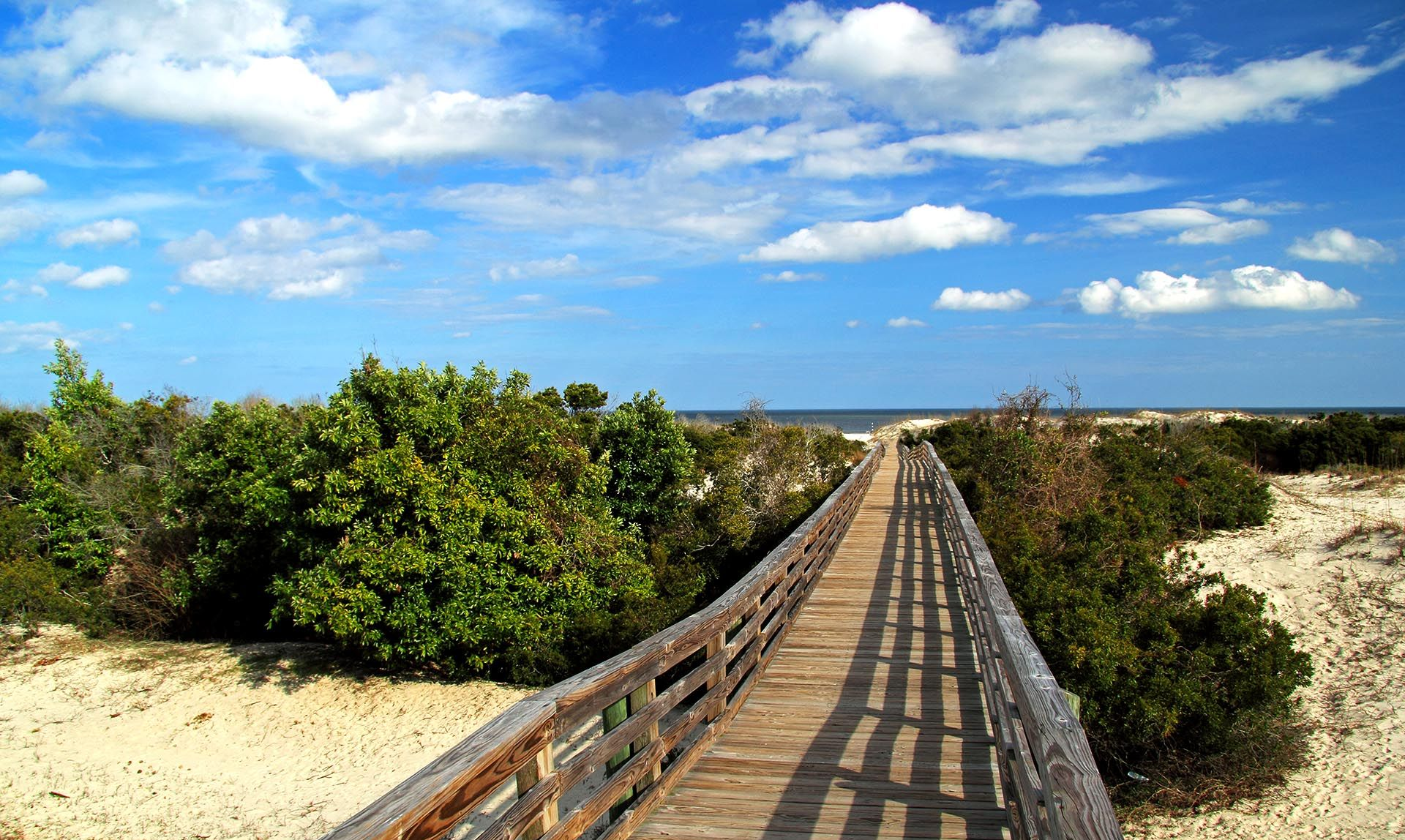 Wooden boardwalk through sandy dunes under a blue sky with scattered clouds
