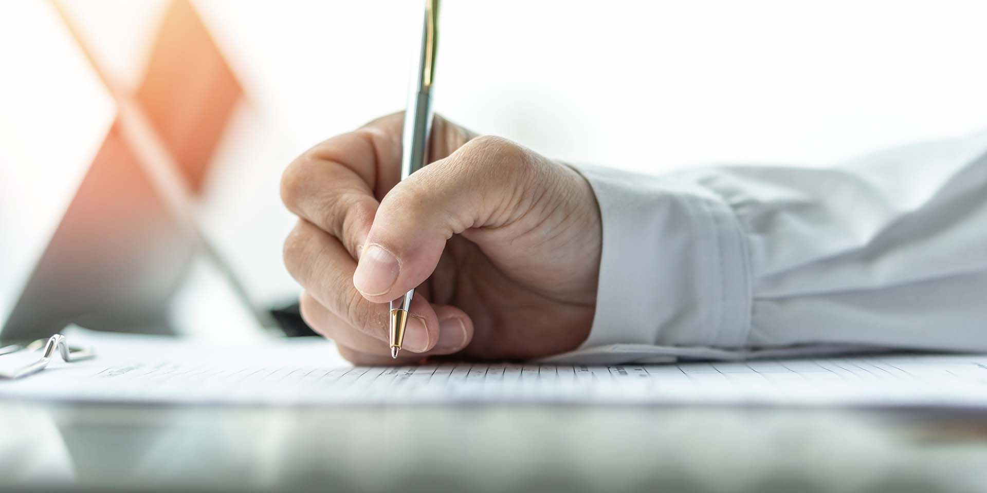 Hand holding a pen over paperwork on a desk beside a laptop and warm light