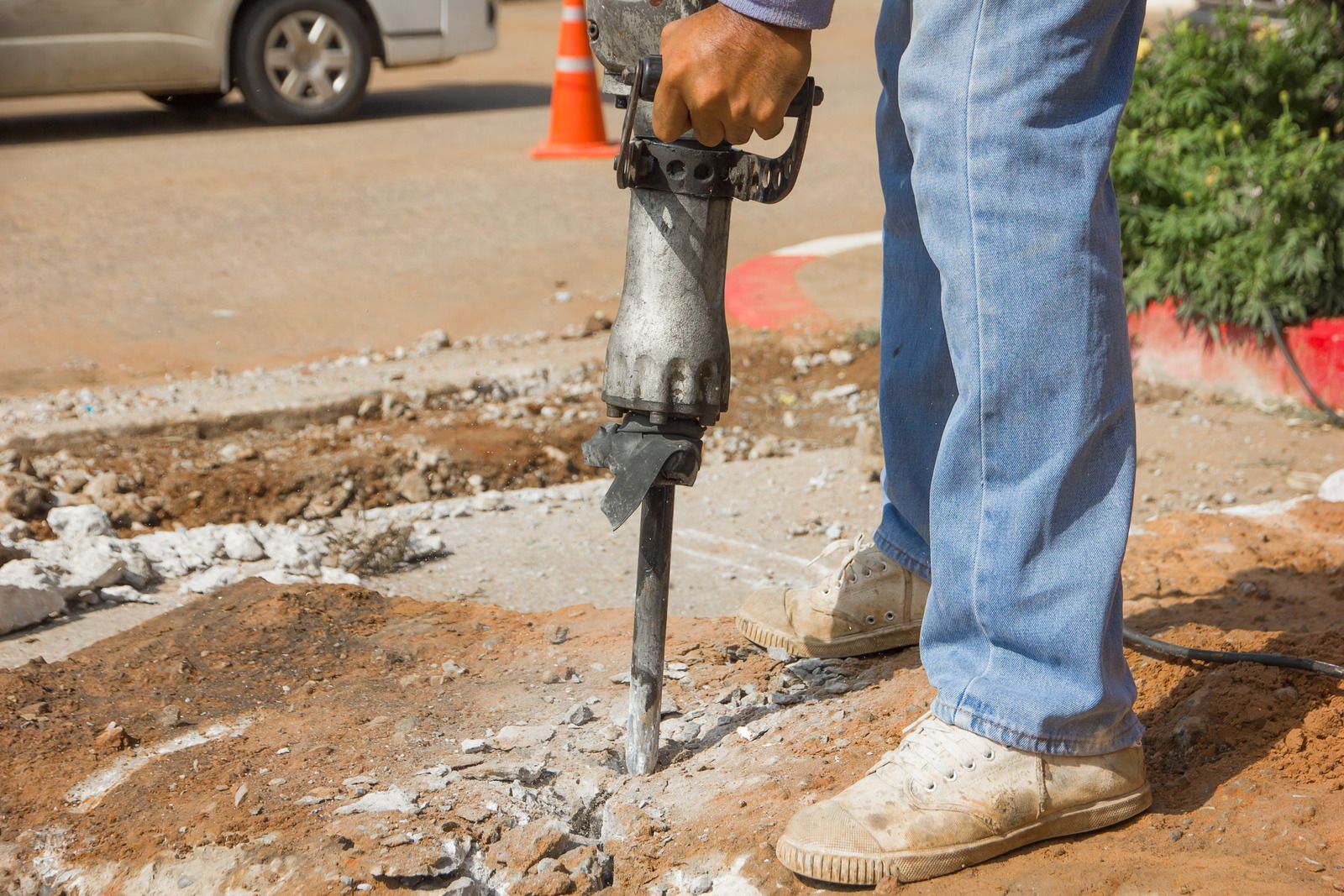 Worker using a jackhammer to break concrete beside a road construction site