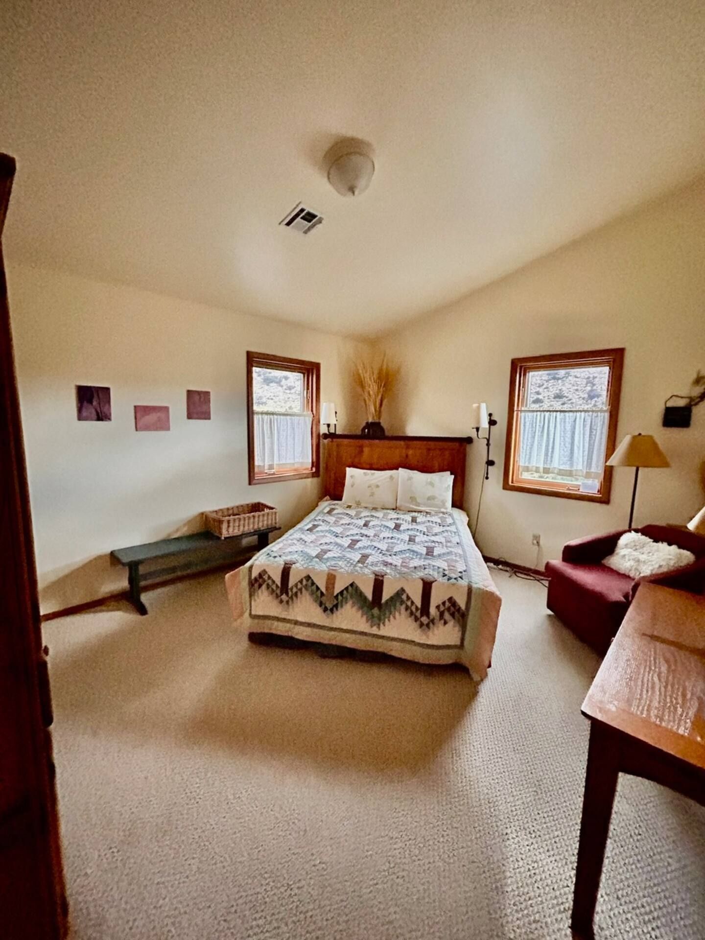 Bedroom with bed, windows, artwork, bench, and armchair. Earth-toned decor, carpeted floor.