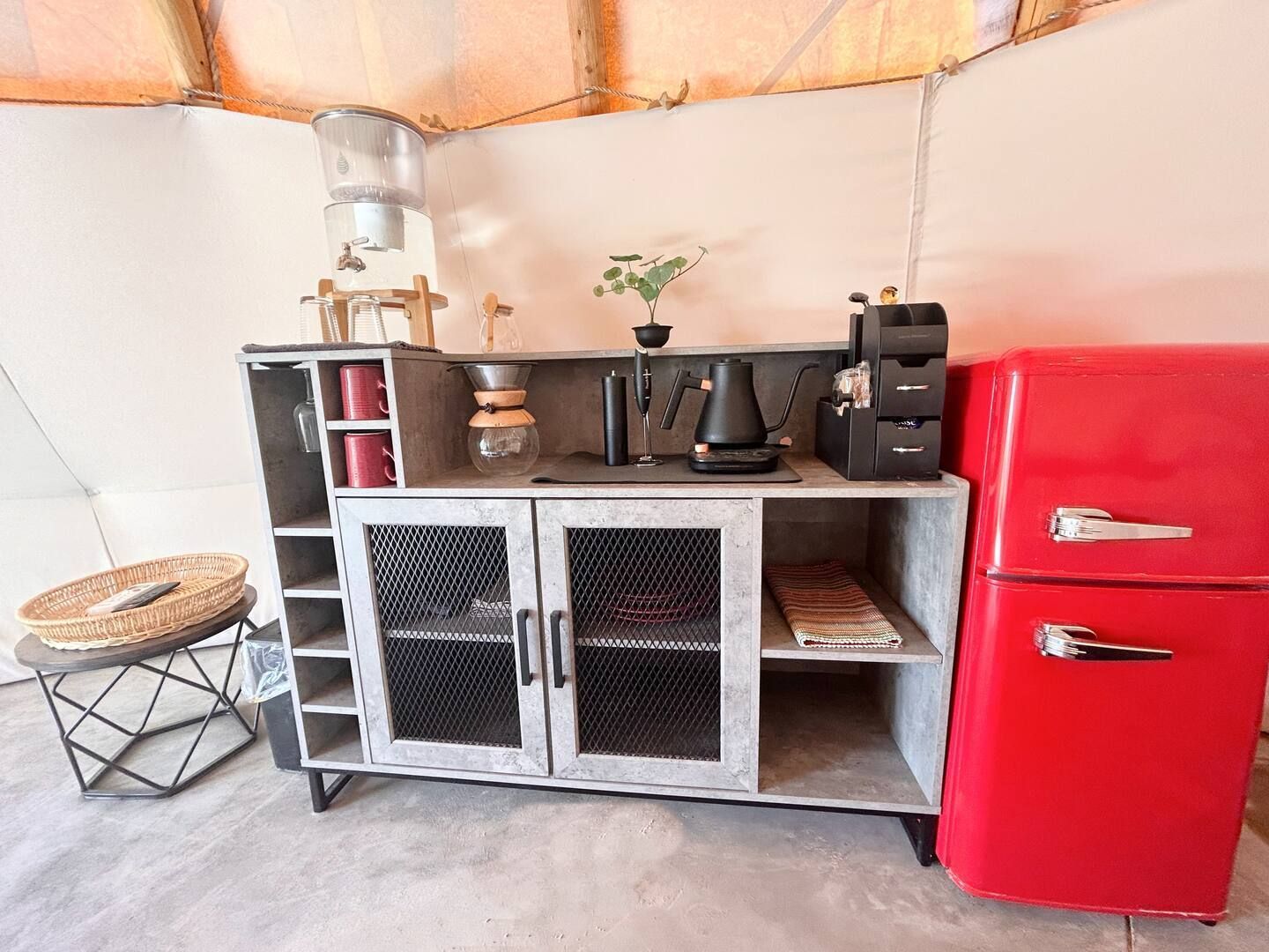 Indoor kitchen area with a red fridge and gray cabinet. Coffee maker, plants, and a small stool present.