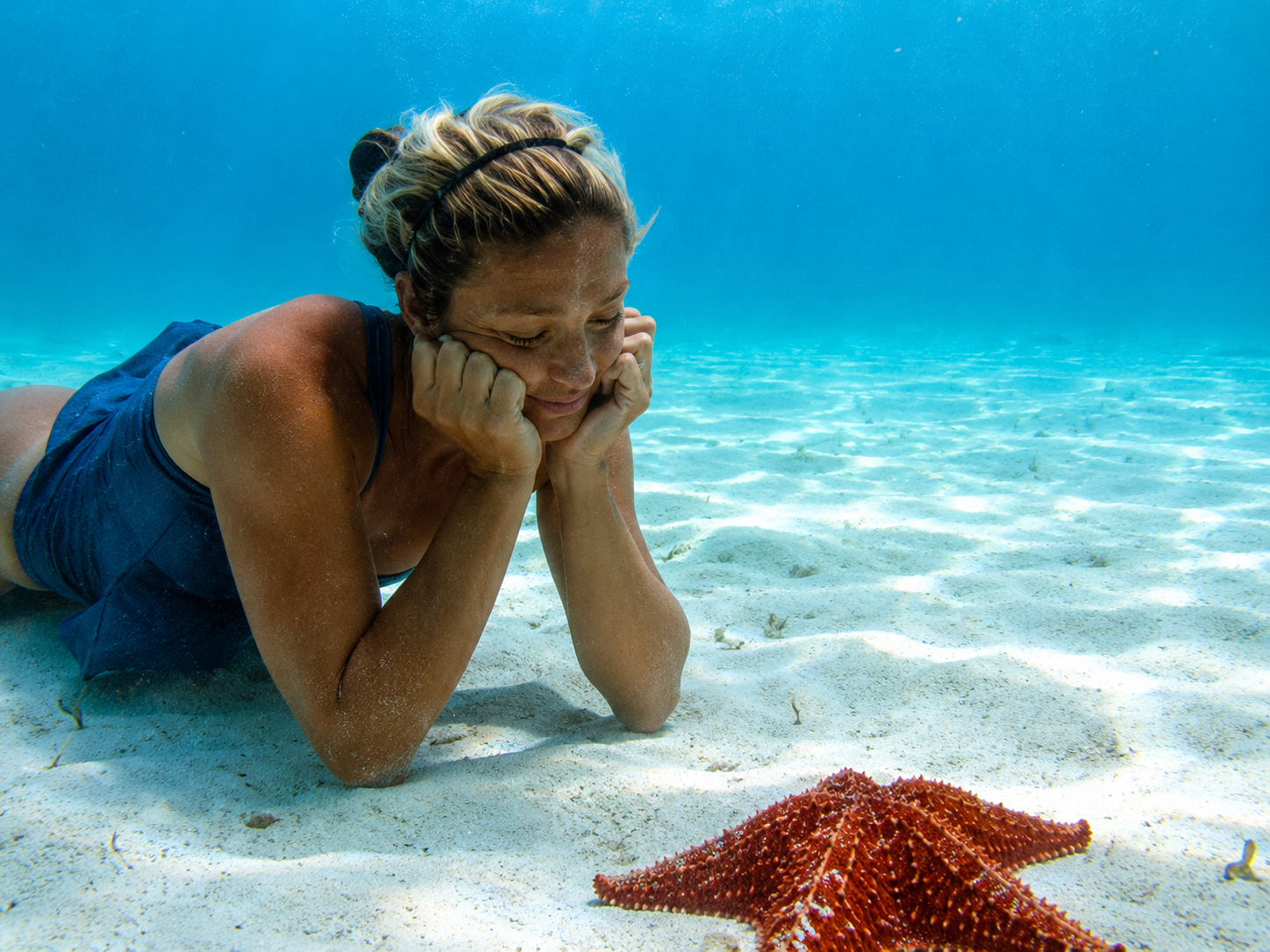 Woman underwater on sandy ocean floor, resting her chin on her hands beside a red starfish.