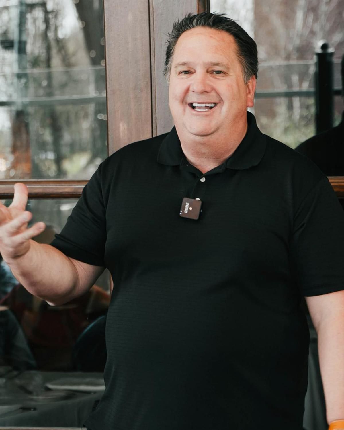 Smiling man in a black polo shirt standing indoors near a window, gesturing with one hand.