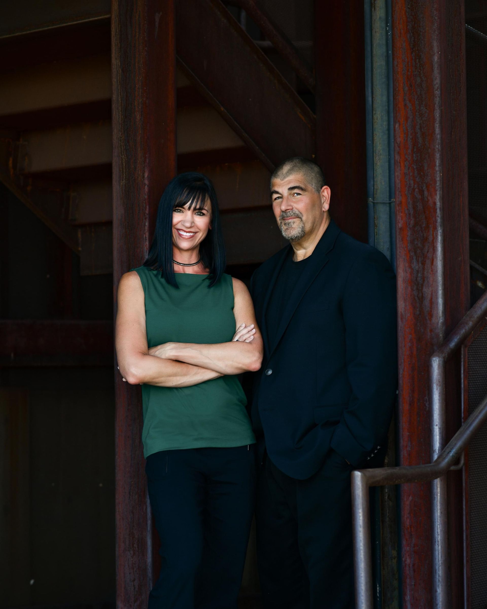 Two people standing with arms crossed in a wooden stairwell, one in green and one in black.