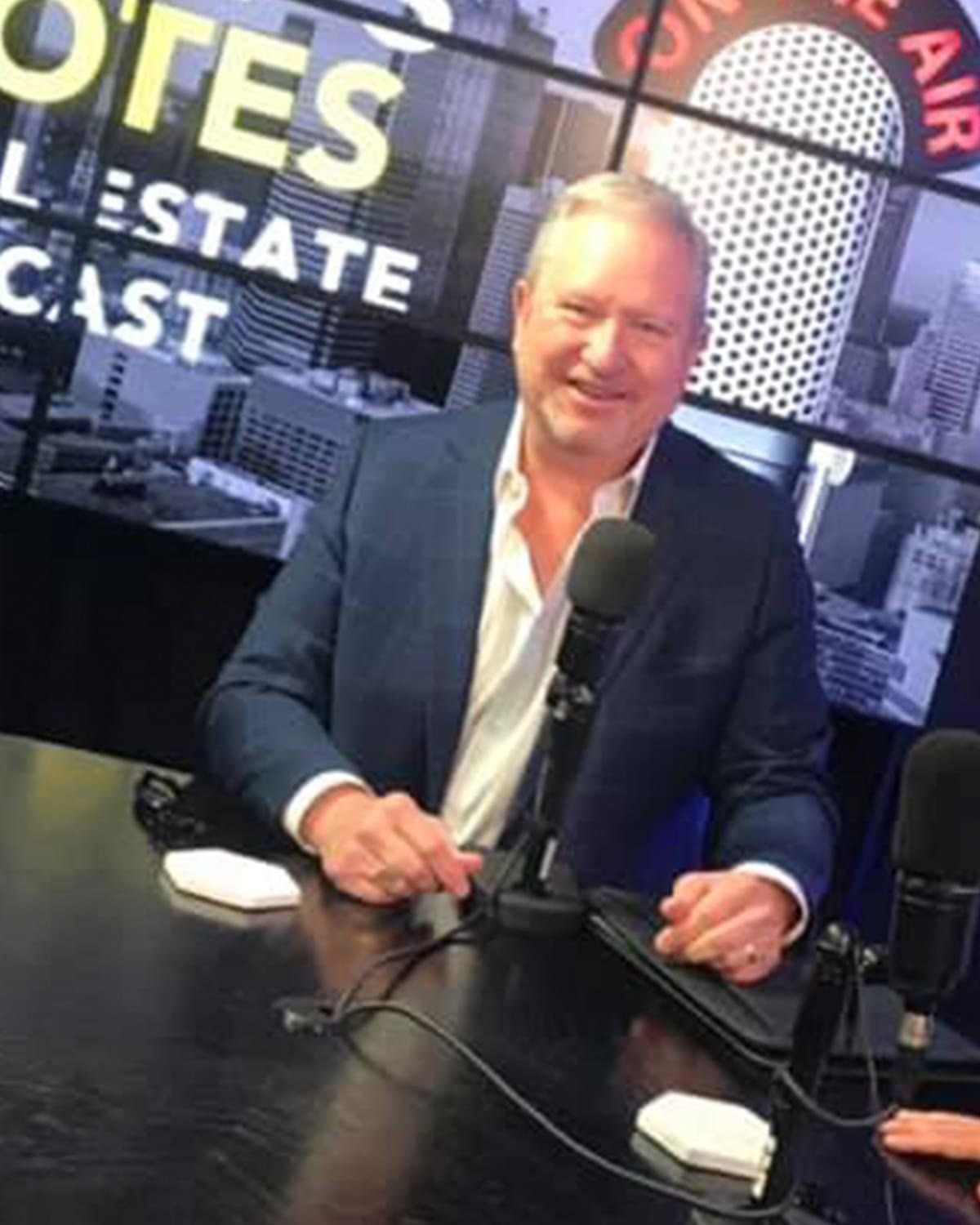 Man in a navy blazer speaking into a microphone at a studio desk with “Real Estate” signage behind him
