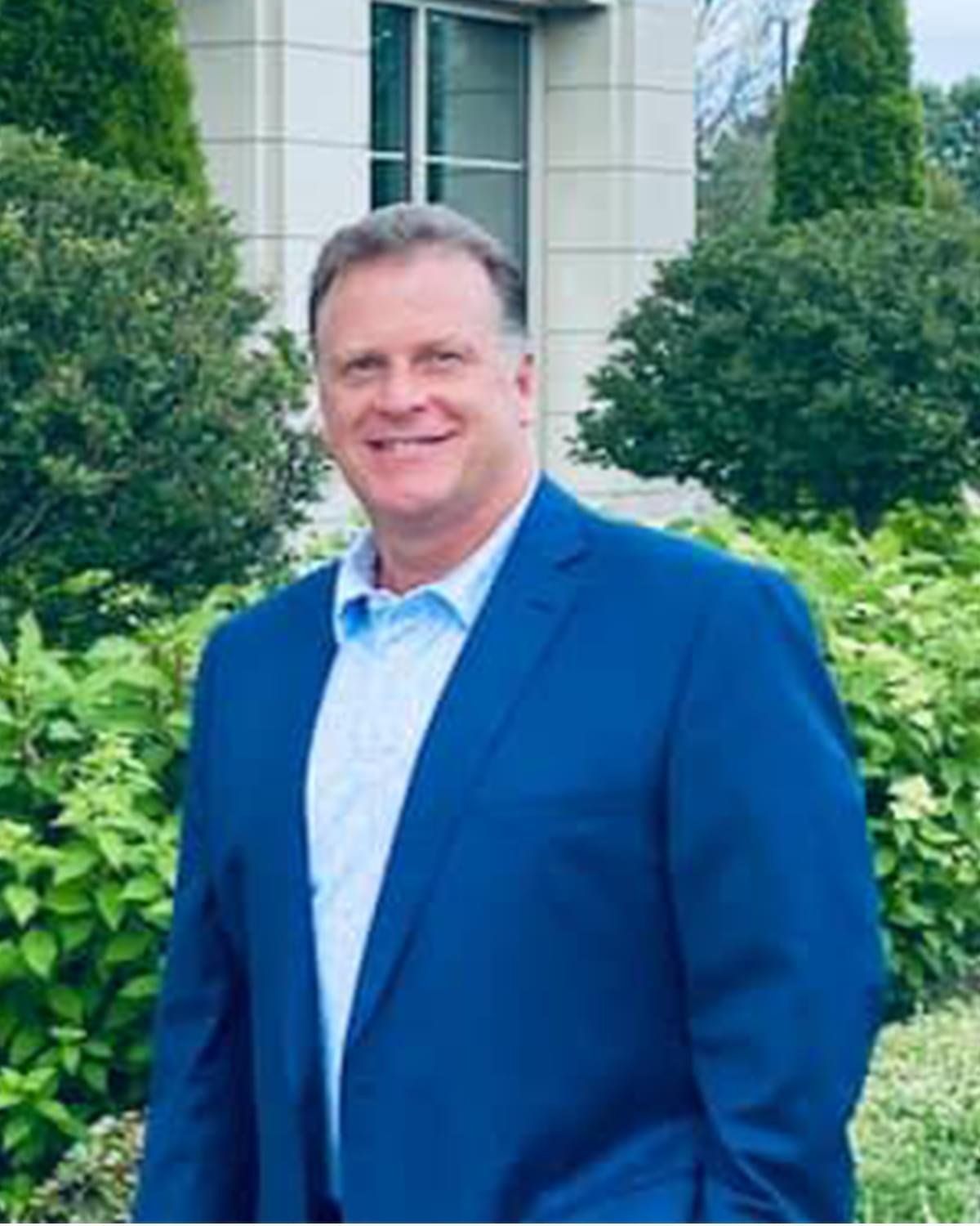 Smiling man in a blue suit jacket standing outdoors in front of greenery and a building