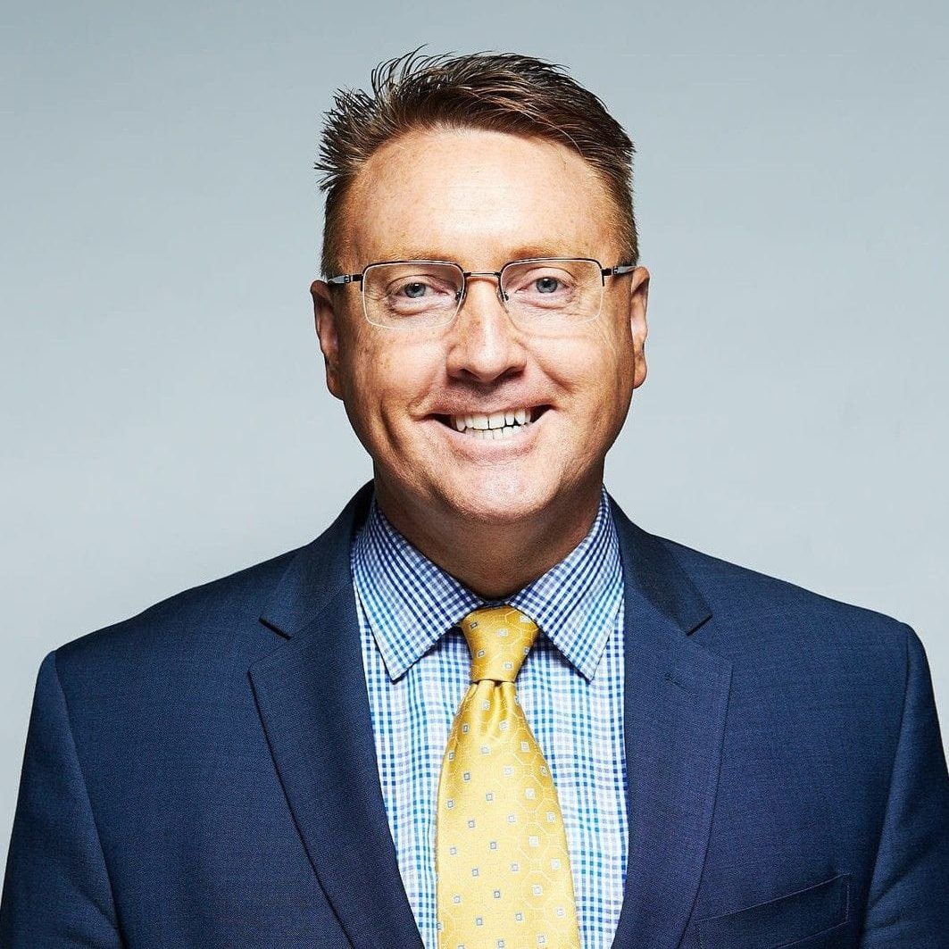 Smiling man in glasses wearing a navy suit, blue checkered shirt, and yellow tie against a gray background