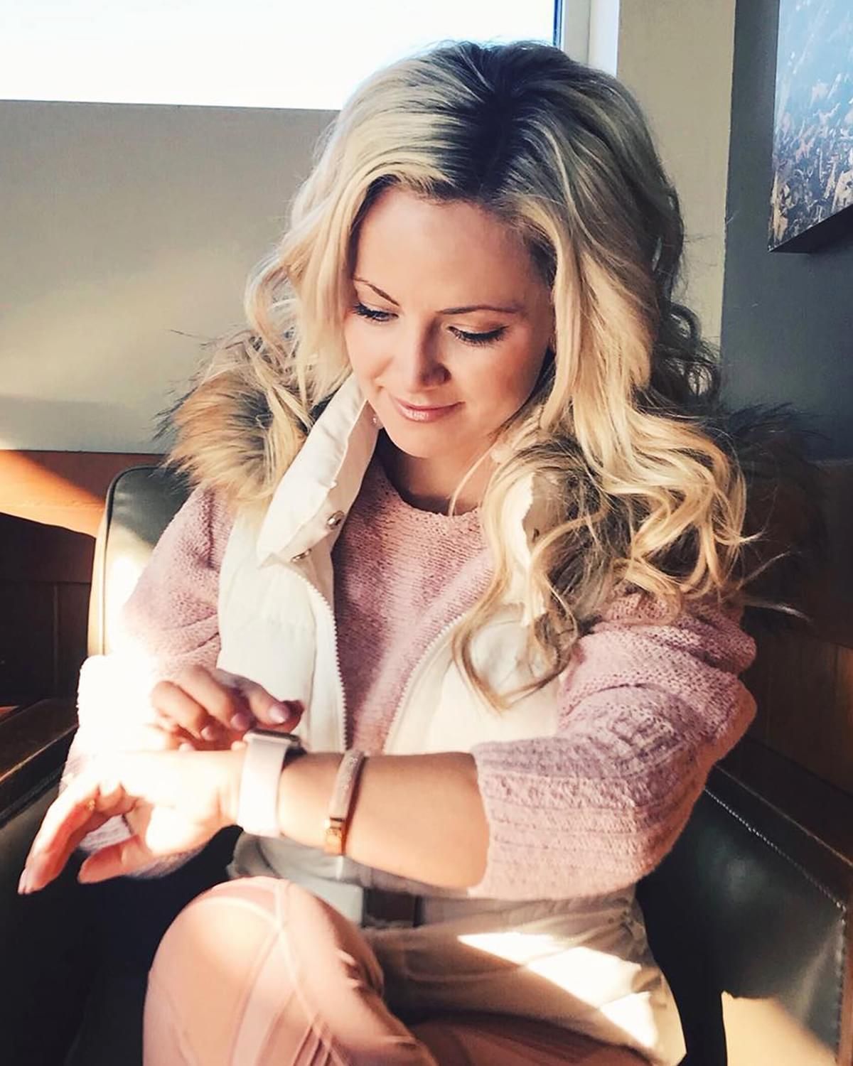 Woman in a pink sweater checking her smartwatch by a sunlit window with a white dog on her lap