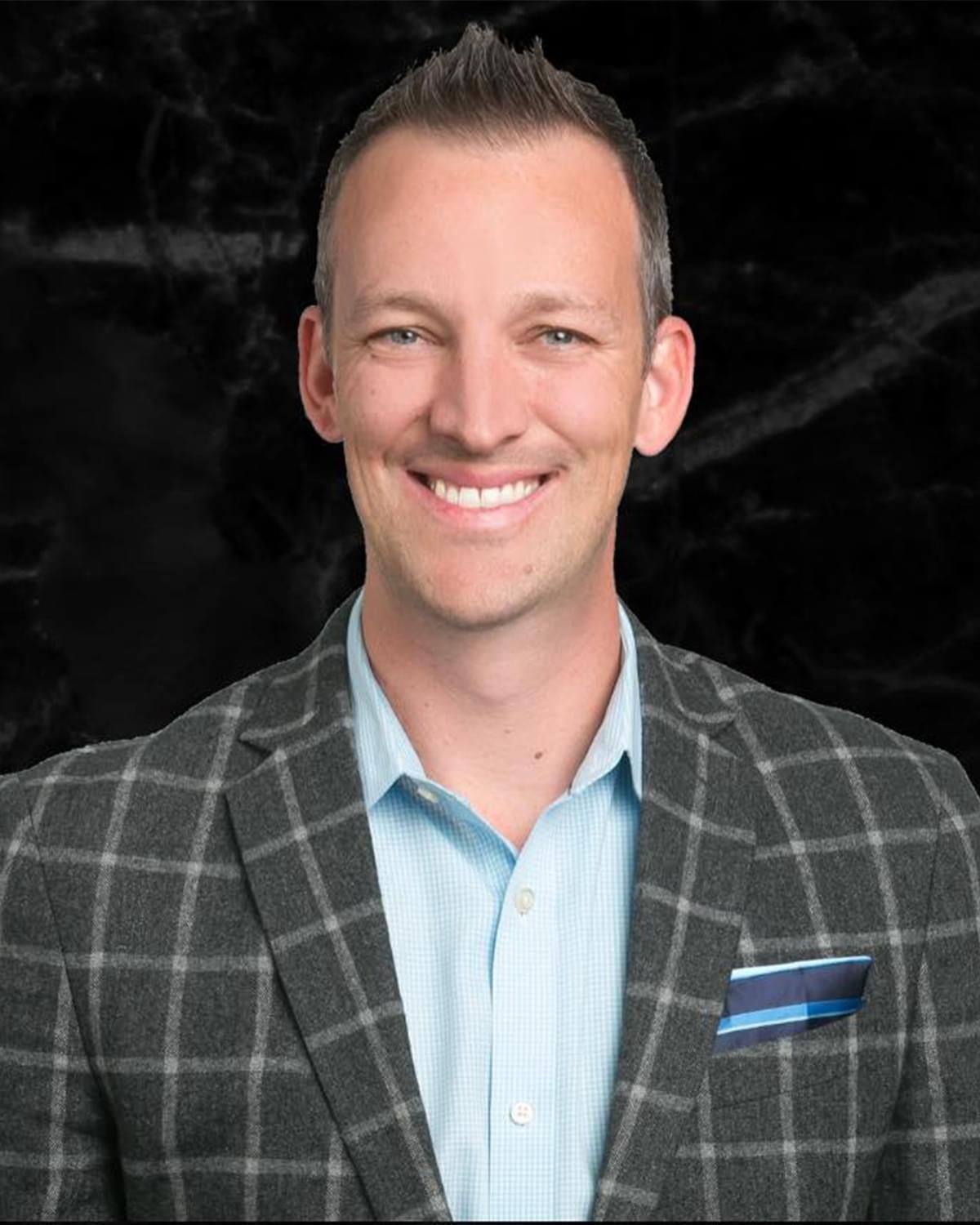 Smiling person in a gray plaid blazer and light blue shirt against a black background.