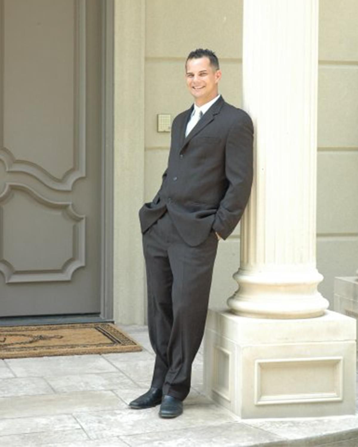 Man in a dark suit leaning against a white column outside a building entrance