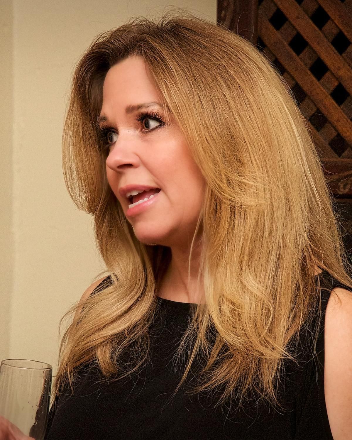 Woman with long blonde hair speaking indoors, wearing a black sleeveless top.