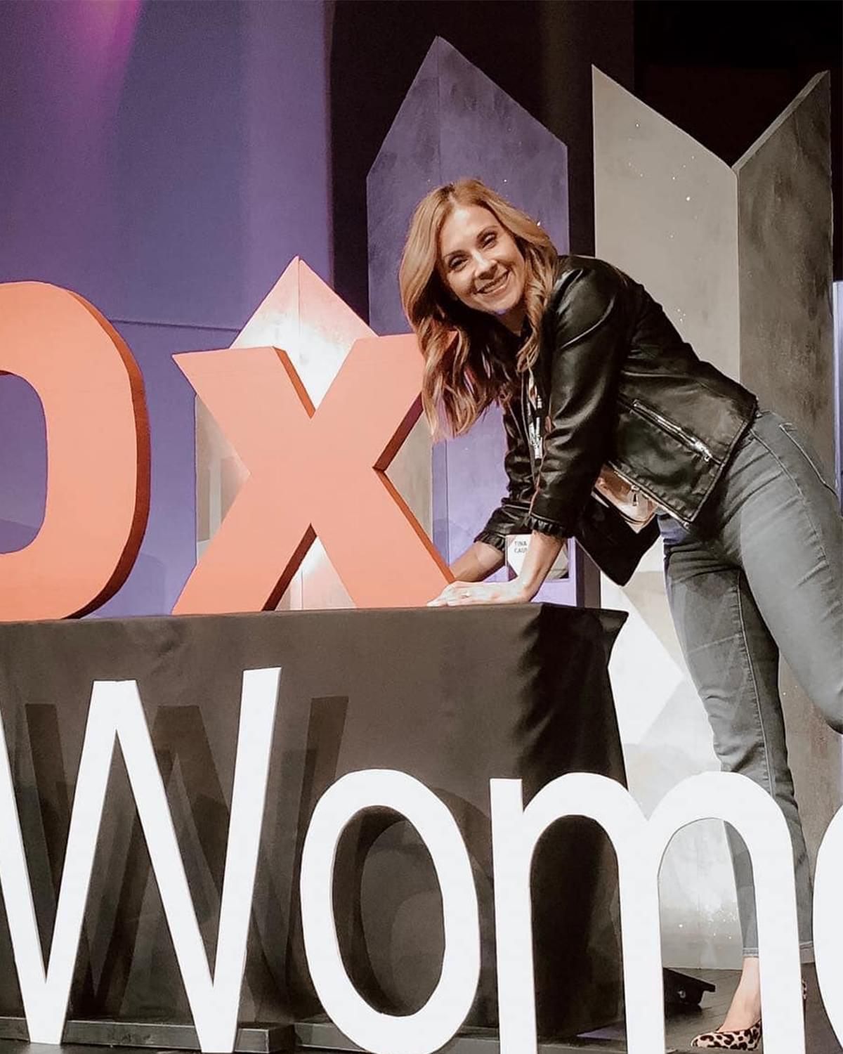 Speaker leaning on TEDx Women stage sign, smiling beside large orange letters on a purple-lit stage