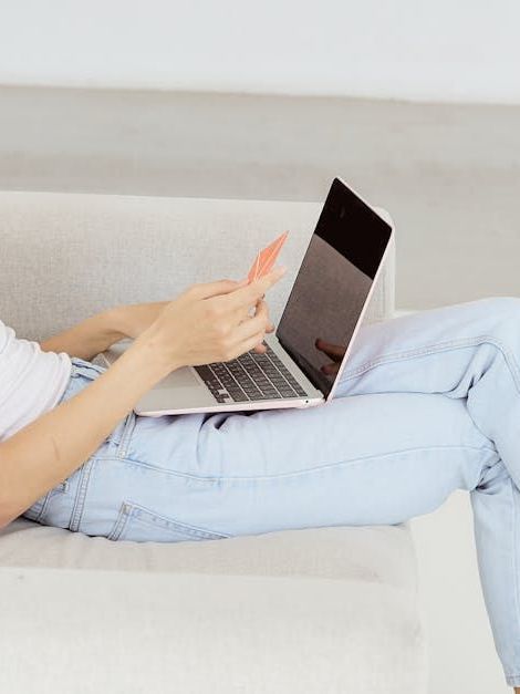 A woman is sitting on a couch using a laptop and holding a credit card.