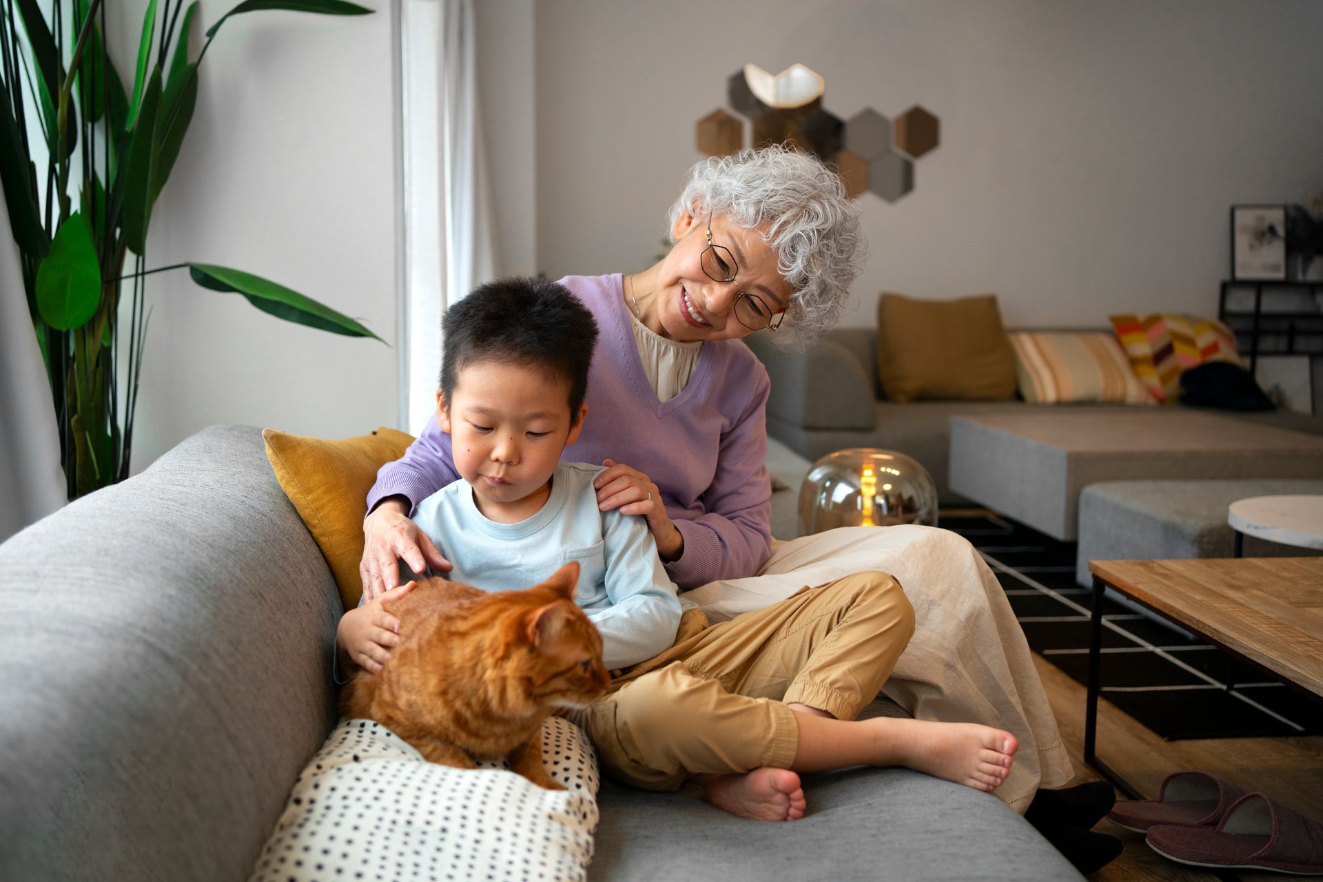 A woman and a young child petting an orange cat on a couch in a living room.
