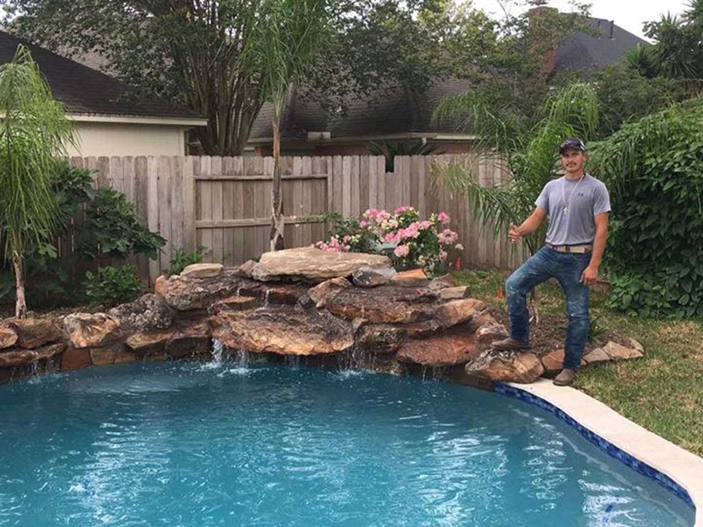 A man is standing next to a swimming pool with a waterfall in the backyard.