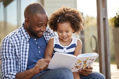 A parent and child sit together outdoors, smiling as they look at an illustrated book.