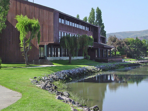A multi-story brown wooden building set beside a tranquil lake with a rocky shoreline and green lawn.