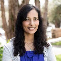 A person with long dark hair, wearing a blue shirt and light-colored cardigan, smiles in an outdoor, wooded setting.
