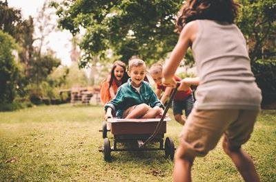 A group of children play in a grassy yard, with one person pulling a red wagon carrying a happy child.
