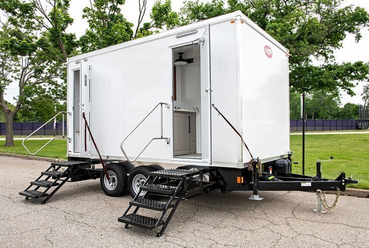 White portable restroom trailer with stairs and handrails parked on asphalt near a grassy field