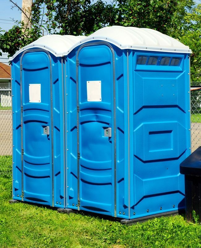 Two blue portable toilets side by side on grass outdoors, with white roofs and a chain-link fence behind them