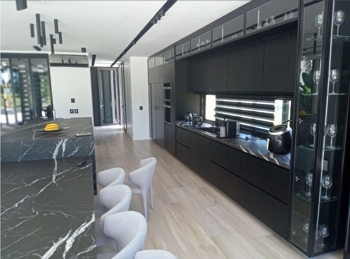 Cocina moderna con isla de mármol negro, armarios negros brillantes y sillas blancas en un interior luminoso.