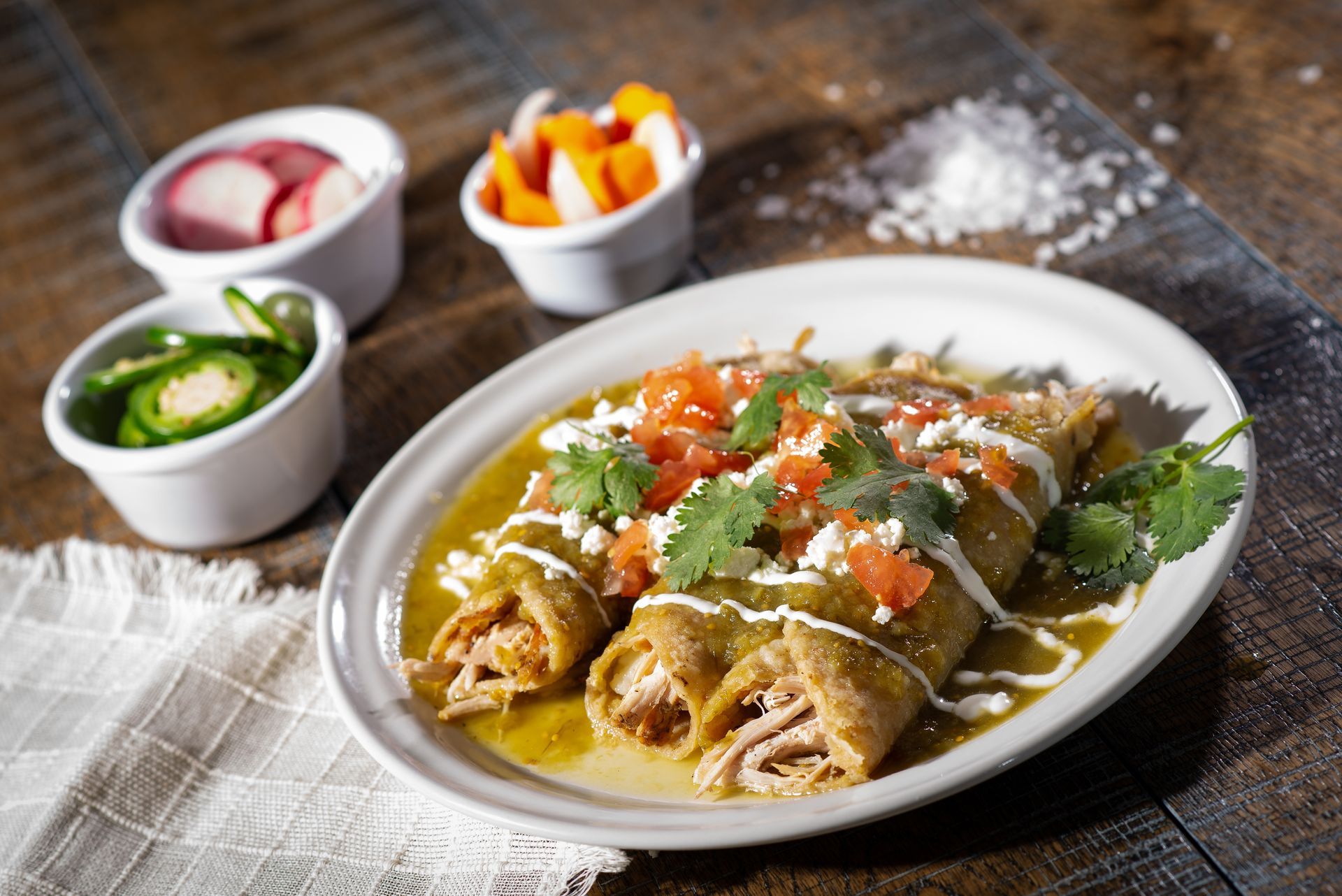 Green enchiladas on a plate, topped with salsa, cheese, and cilantro, with garnishes and salt.