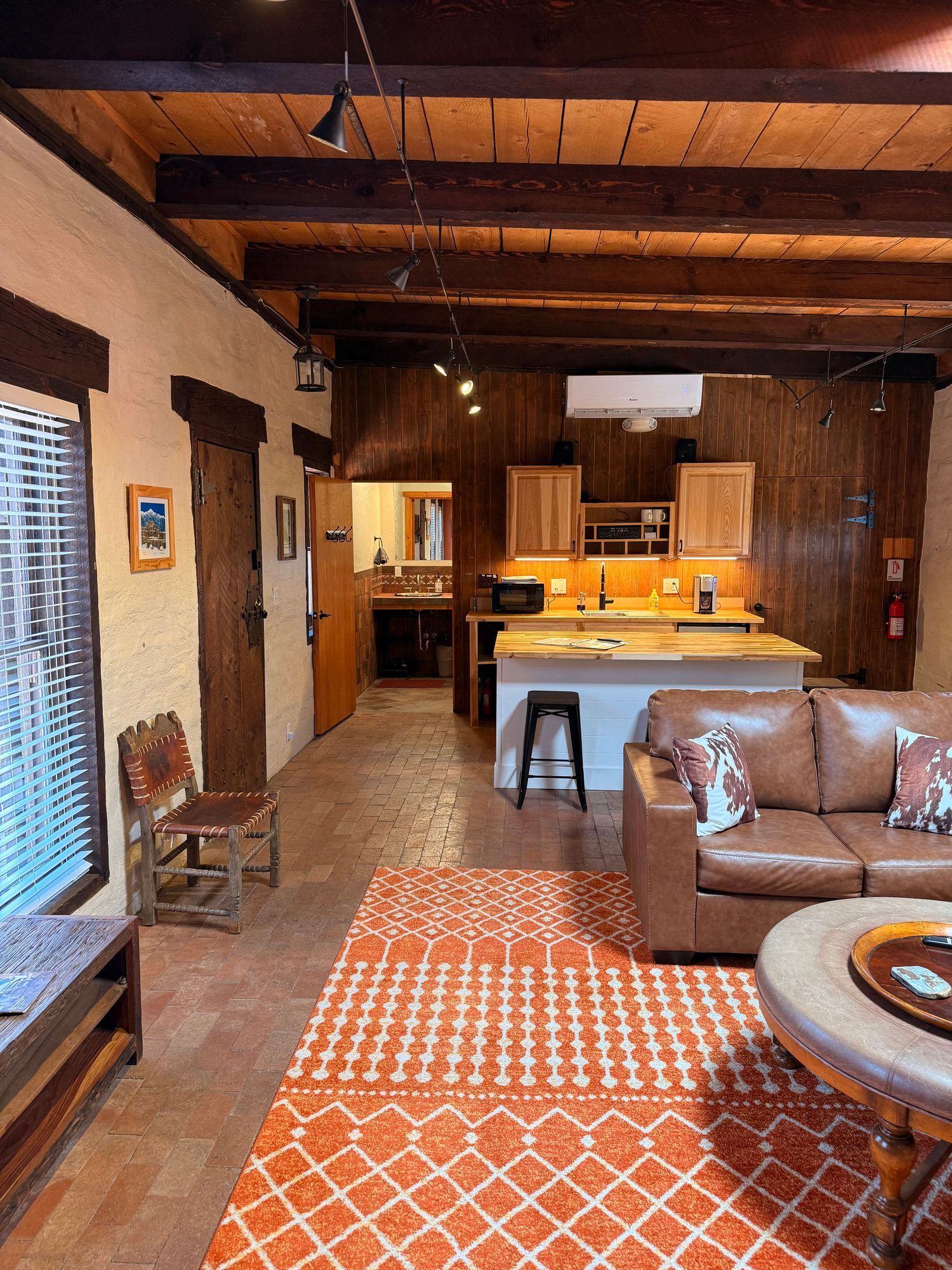 Cozy living area with leather sofa, orange rug, open kitchen, and rustic wooden beams.