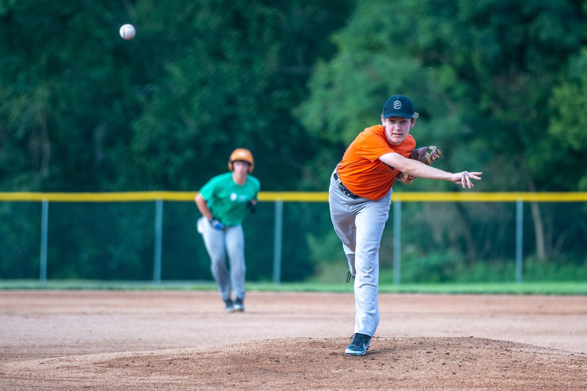 Baseball pitcher in orange throws on a dirt field, with a fielder in green behind him.
