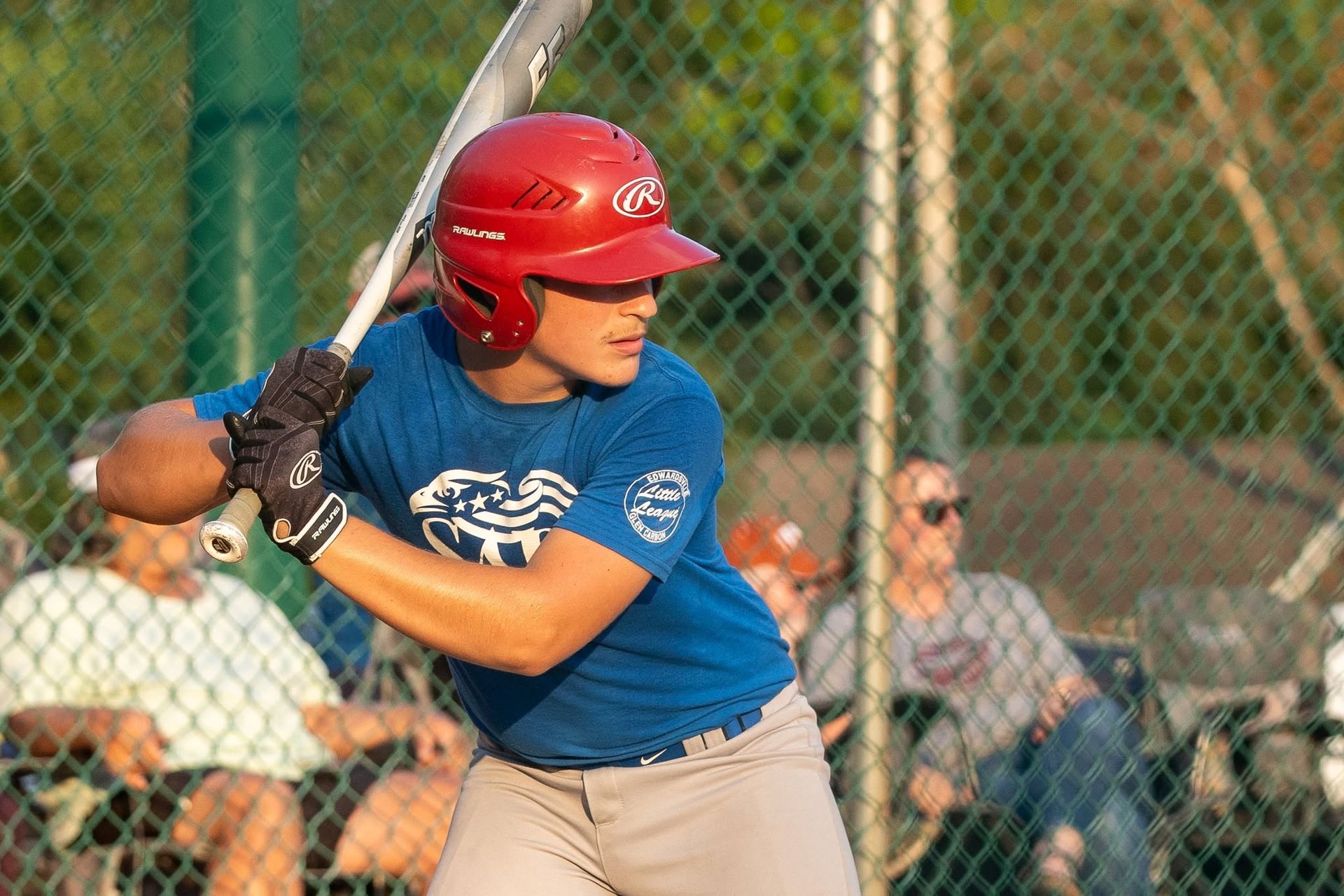 Youth softball player in blue jersey and red helmet batting at the plate during a game.