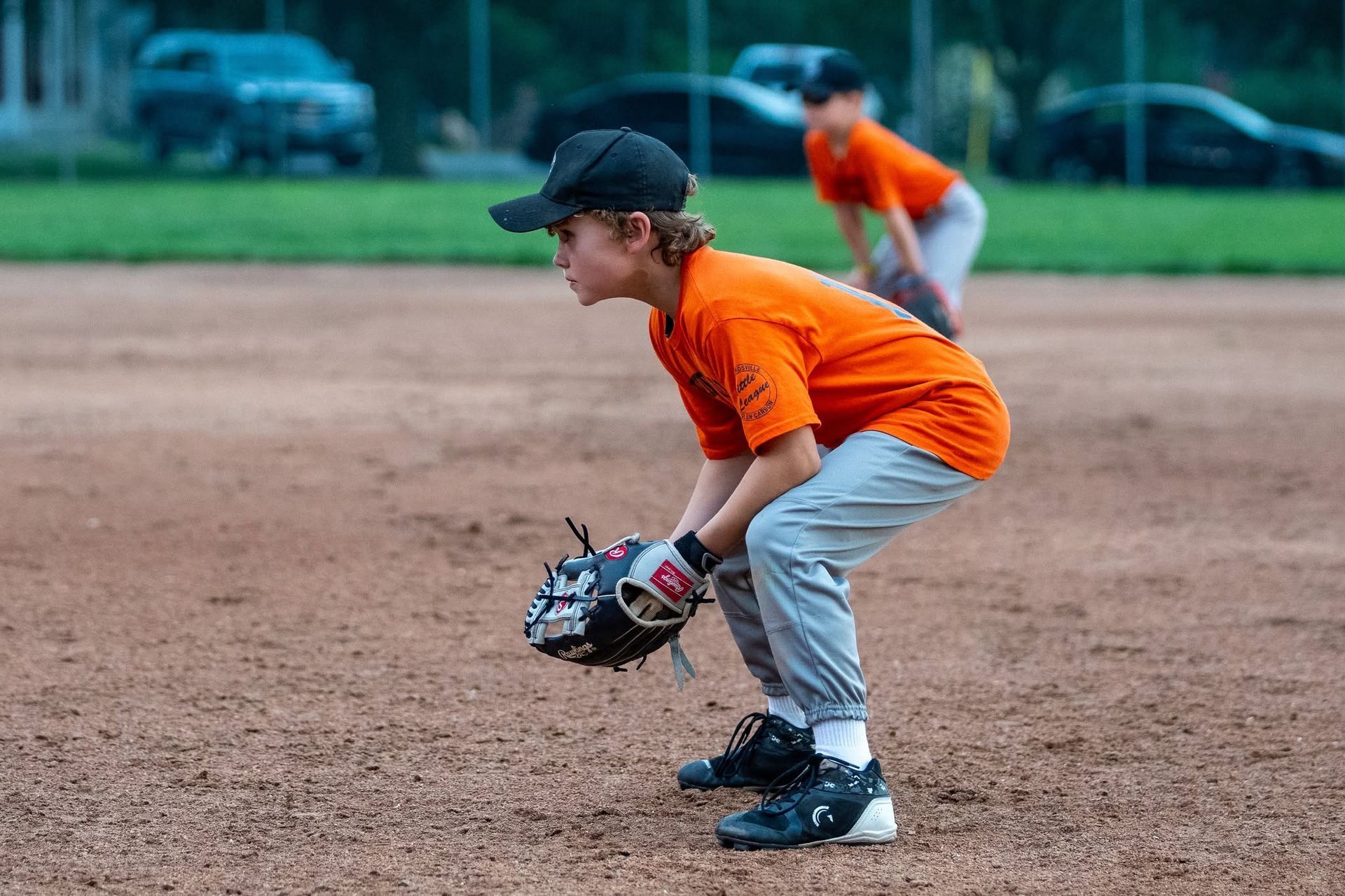 Youth baseball player in orange jersey crouches on a dirt field, ready to catch a ball.