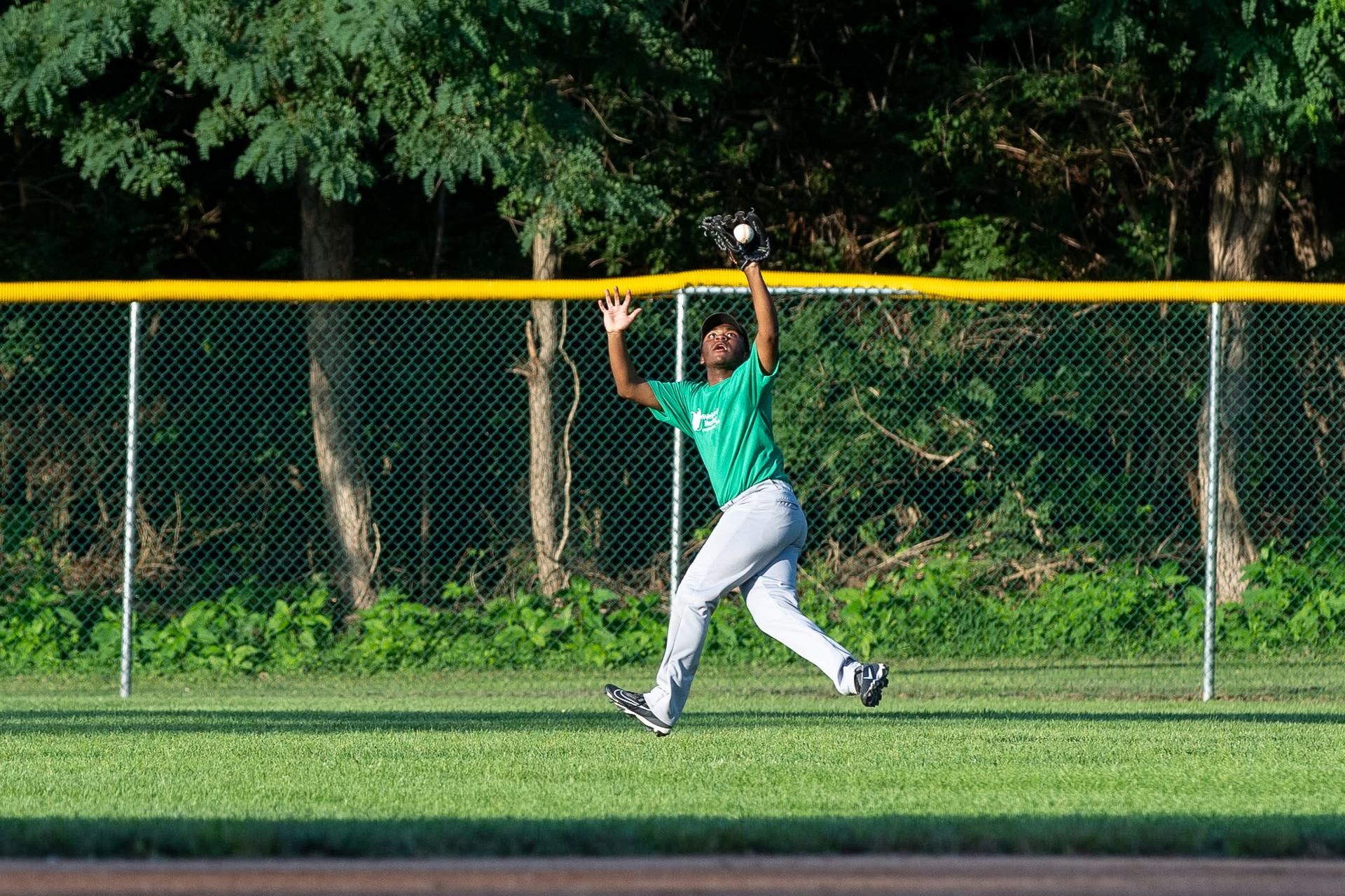 Person in green shirt leaps to catch a ball near a yellow fence on a sports field.