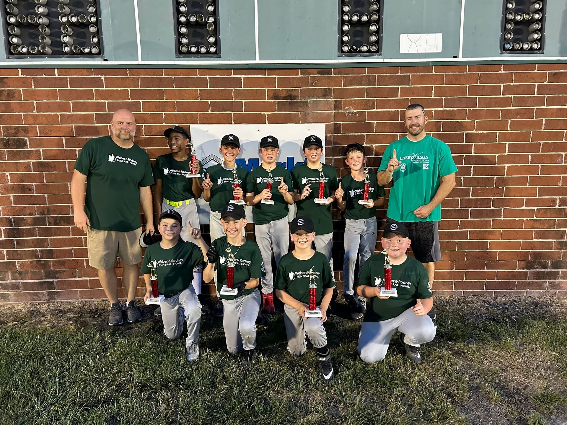 Youth baseball team posing with coaches in front of a brick wall and scoreboard.