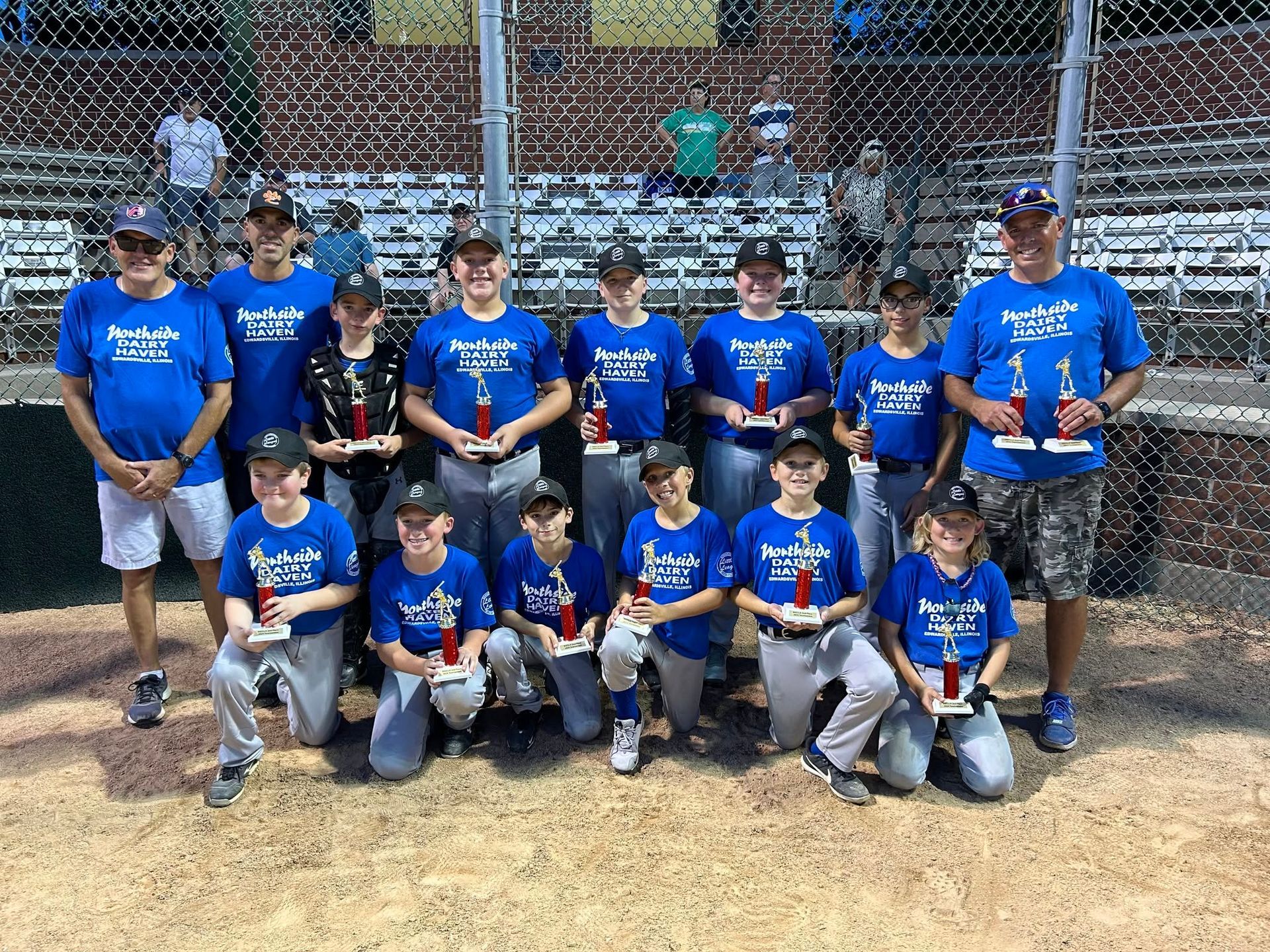 Youth baseball team in blue shirts posing on a field in front of a chain-link fence with bleachers behind them