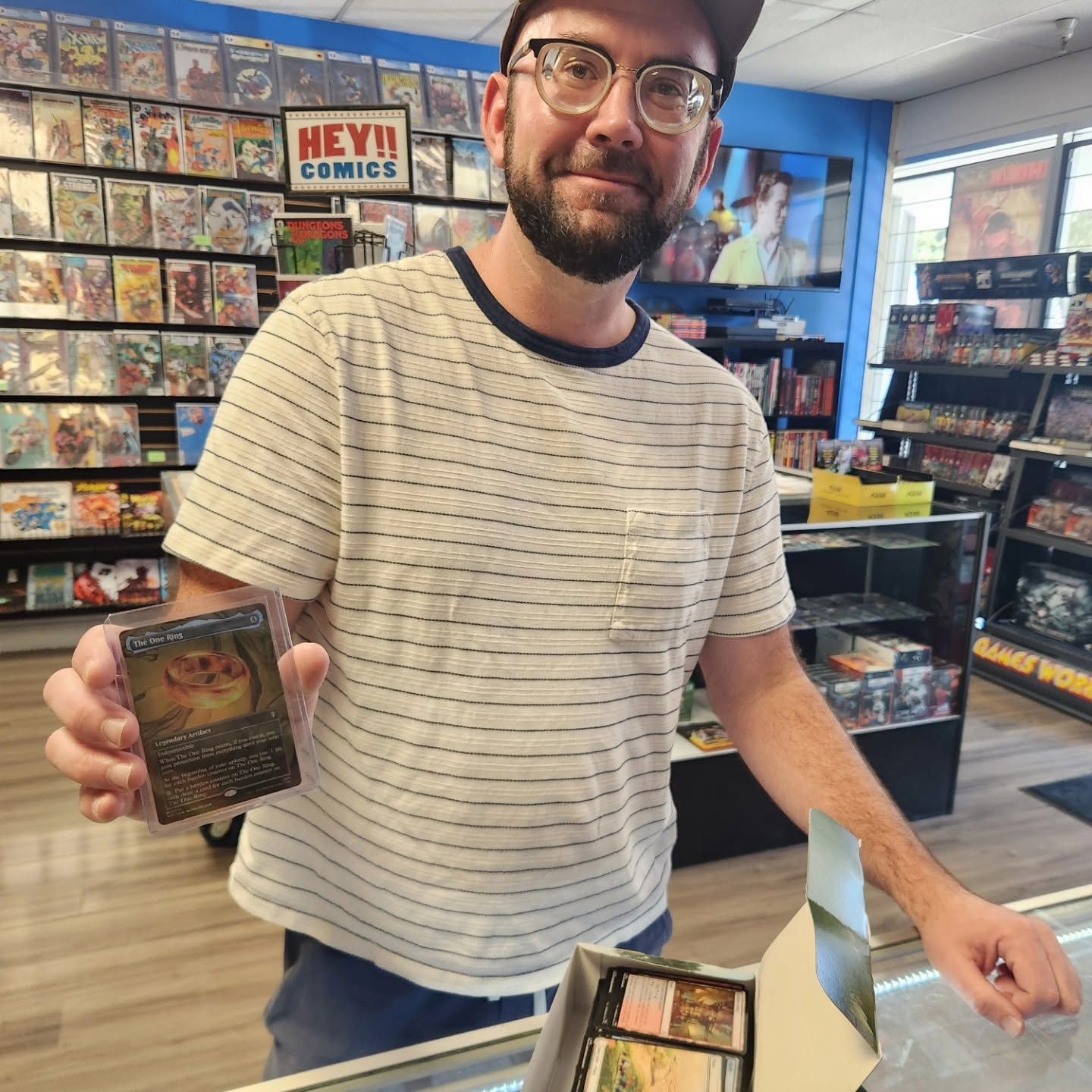 Man holding a trading card in a comic shop, standing by a counter with display boxes.