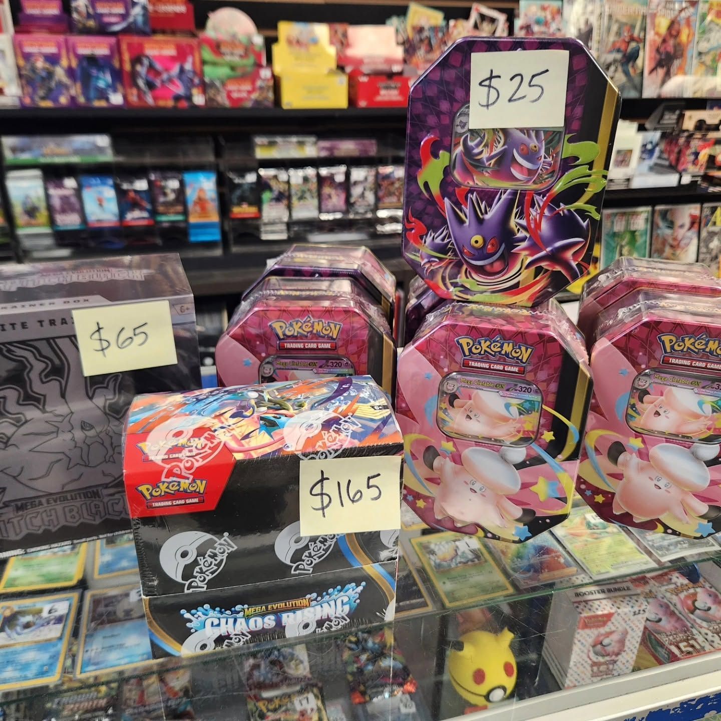 Pokémon trading card tins and boxes stacked in a store display with price tags of $25, $65, and $165