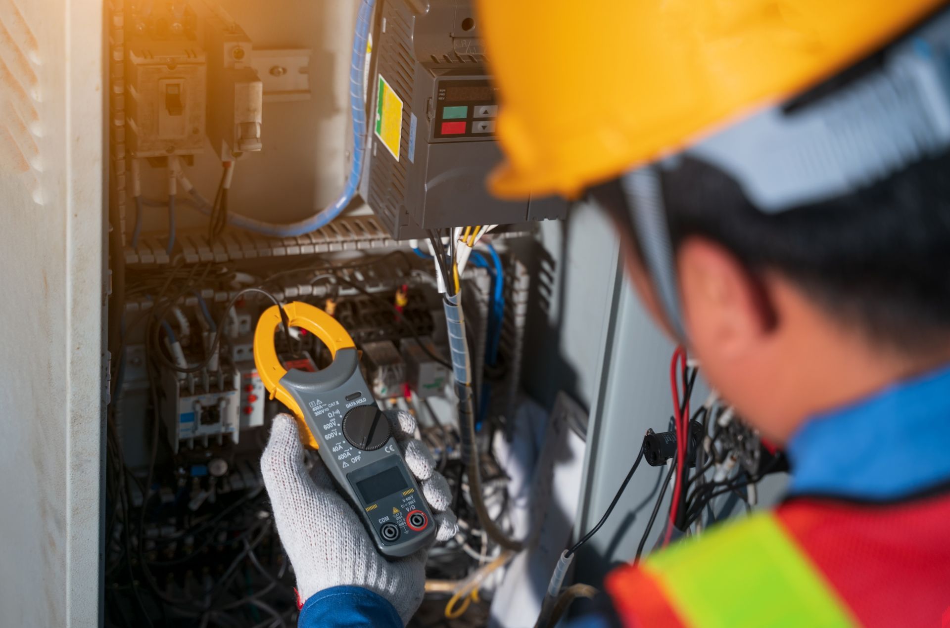 A technician in a hard hat and safety vest uses a clamp meter to test electrical components inside a control panel.